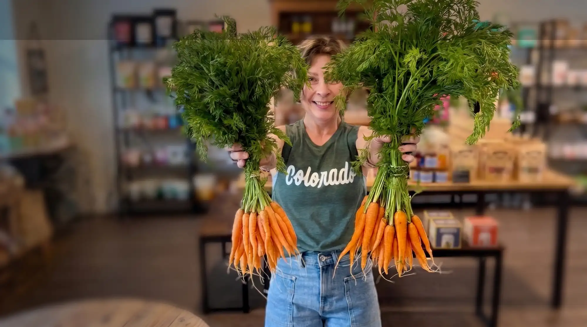 A smiling shopkeeper holds up two big bunches of fresh carrots inside Mountain Market & Supply in Eagle